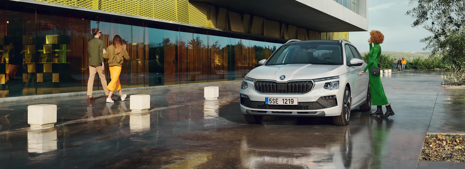 Škoda Kamiq parked in the front of a modern building with a woman entering while a young couple passes by, ¾ front angle, moon white metallic color