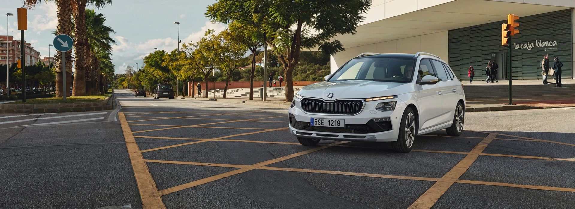 Škoda Kamiq parked in the front of a modern building with a woman entering while a young couple passes by, ¾ front angle, moon white metallic color