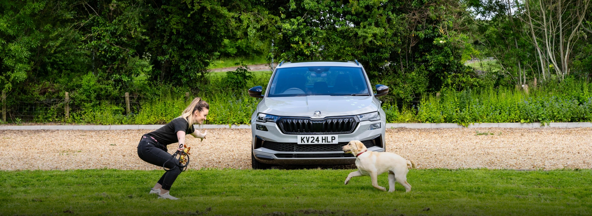 A grey Škoda Karoq parked behind a woman training a Labrador on a green lawn.