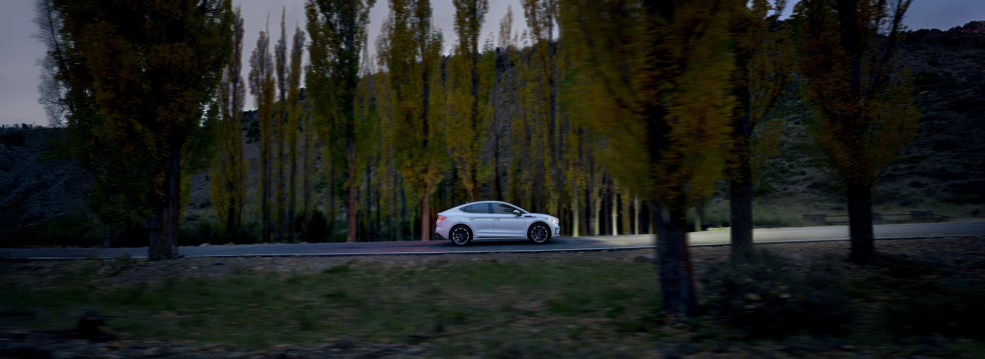 White Škoda saloon driving on a tree-lined road at dusk; a safety-focused hero image.