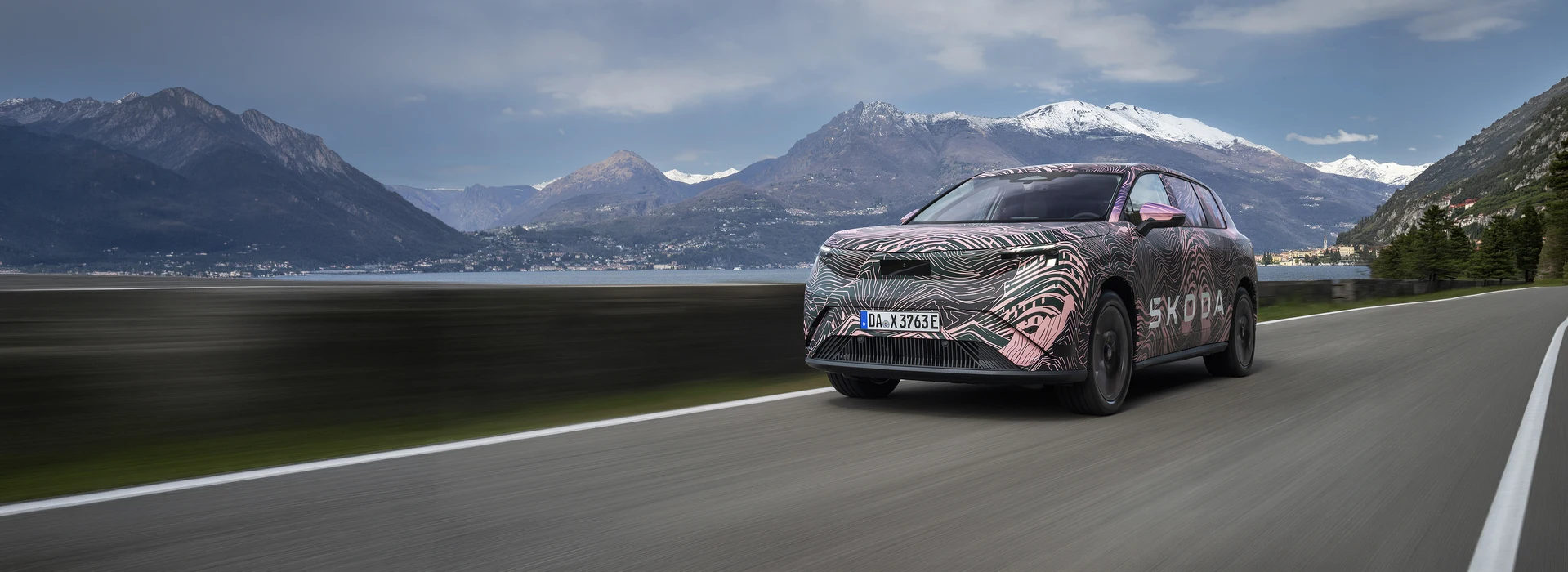 Front three-quarter view of a Škoda Peaq in pink camouflage driving alongside a scenic lake.