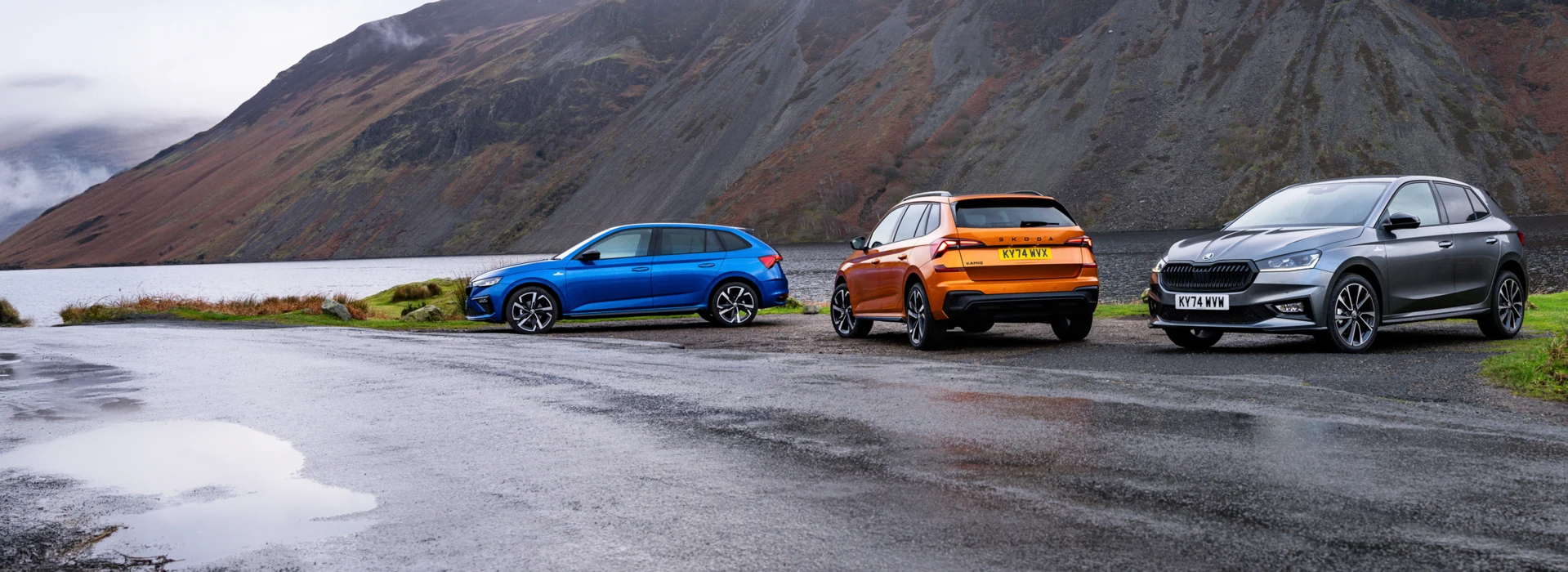 Blue, orange and grey Škoda cars parked on a wet mountain road under cloudy skies.