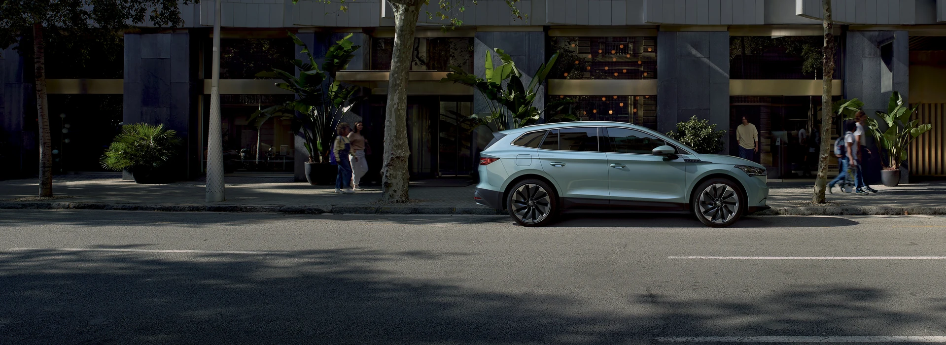 A photo of a crystal blue ENYAQ parked on the side of the street with people walking in the sidewalk. A grey building is seen in the background