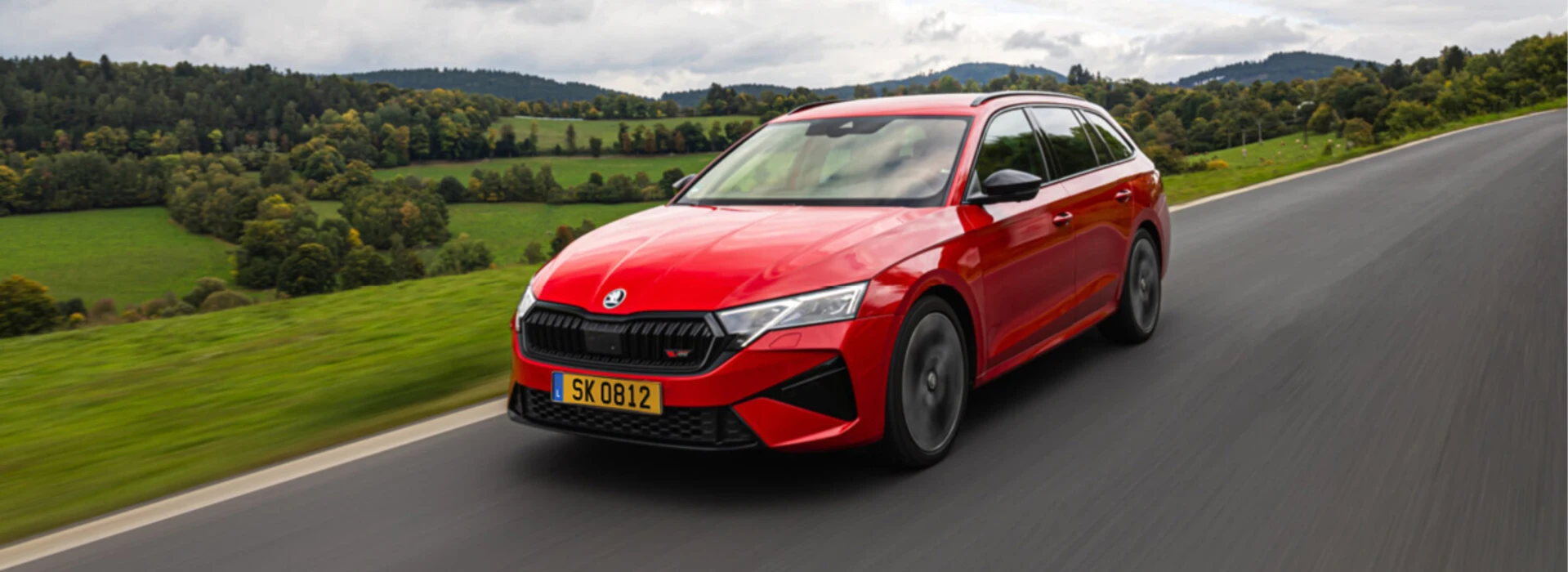 Une Škoda Octavia Combi rouge circulant sur une route de campagne bordée de collines verdoyantes.