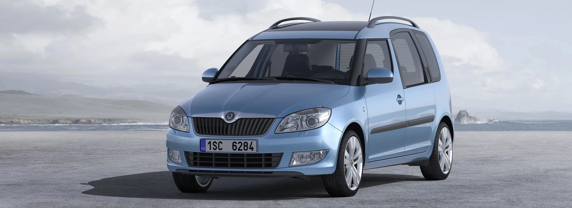 Une voiture de couleur bleu clair métallisé, de modèle Škoda Roomster, stationnée sur une surface plane avec un paysage côtier et un ciel nuageux en arrière-plan.