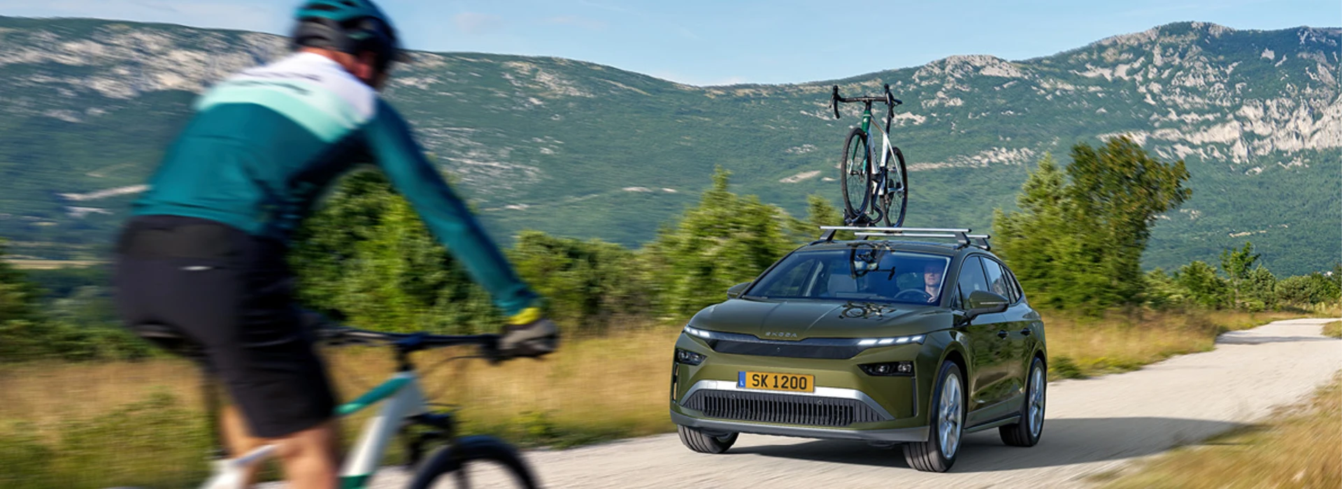 Une voiture Skoda, probablement un SUV, de couleur foncée, est garée sur un chemin de terre en montagne. Au premier plan et sur la gauche, on voit une personne avec un vélo tout-terrain (VTT) ou de montagne, vêtue d'une tenue de cycliste.