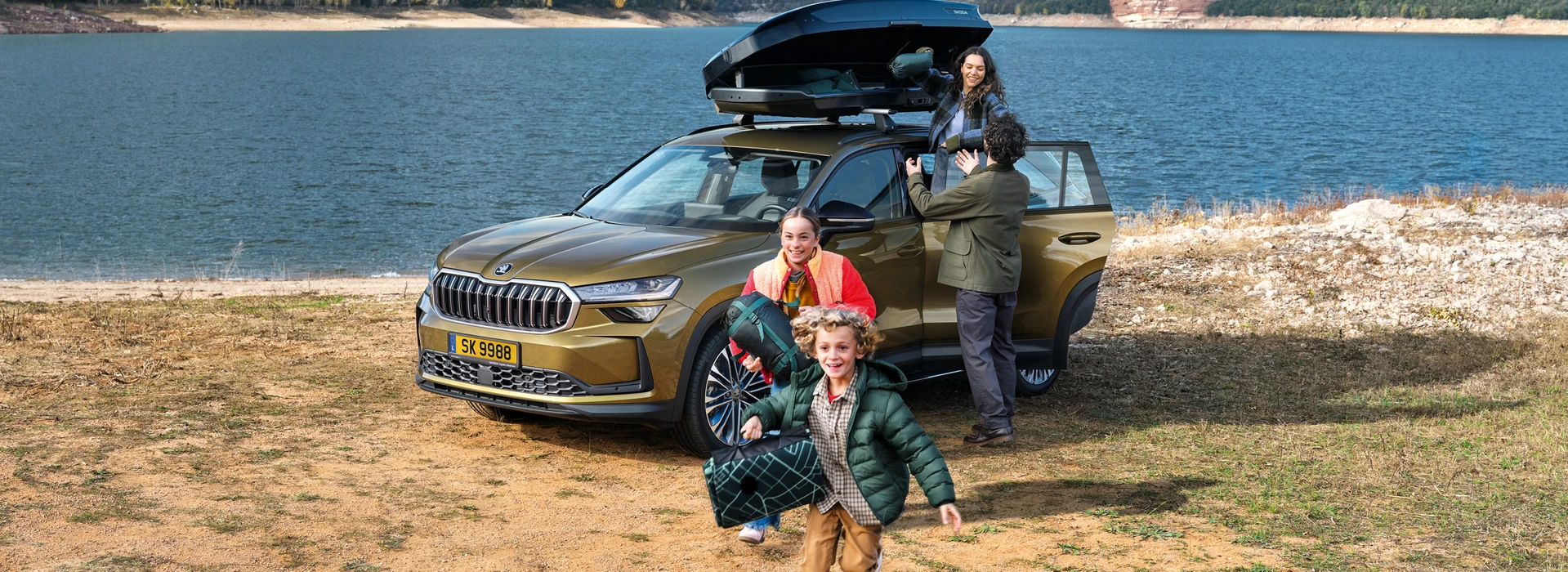 Famille chargeant une voiture Škoda près d’un lac, avec des enfants jouant et des bagages sur le toit.