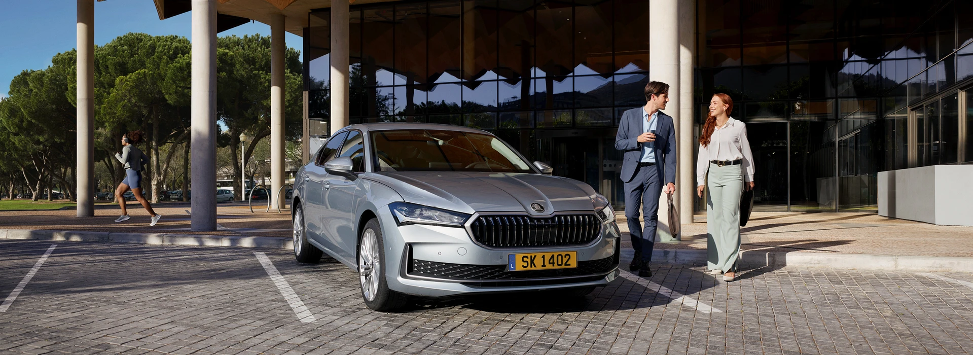 Une Skoda Superb de couleur gris argent est stationnée sur un parking. Deux personnes, un homme et une femme habillés de manière professionnelle, se tiennent à l'avant gauche de la voiture, face à l'entrée d'un bâtiment moderne.