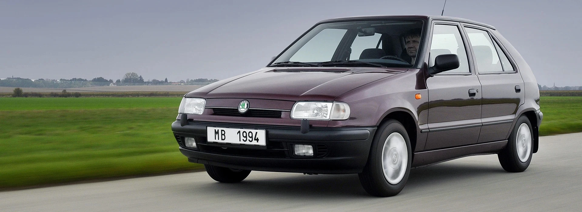 Une voiture compacte Škoda Felicia de couleur bordeaux foncé, circulant sur une route avec un paysage verdoyant en arrière-plan