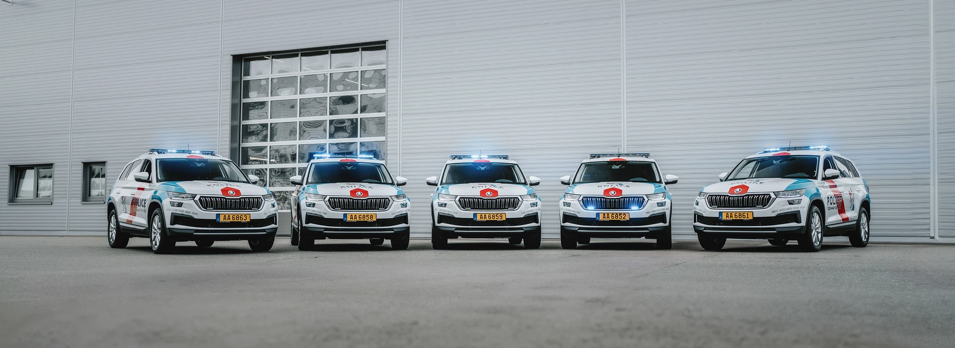 Cinq voitures de police blanches et rouges sont garées en ligne droite, devant un bâtiment gris avec une grande porte de garage. Leurs gyrophares bleus sont allumés.