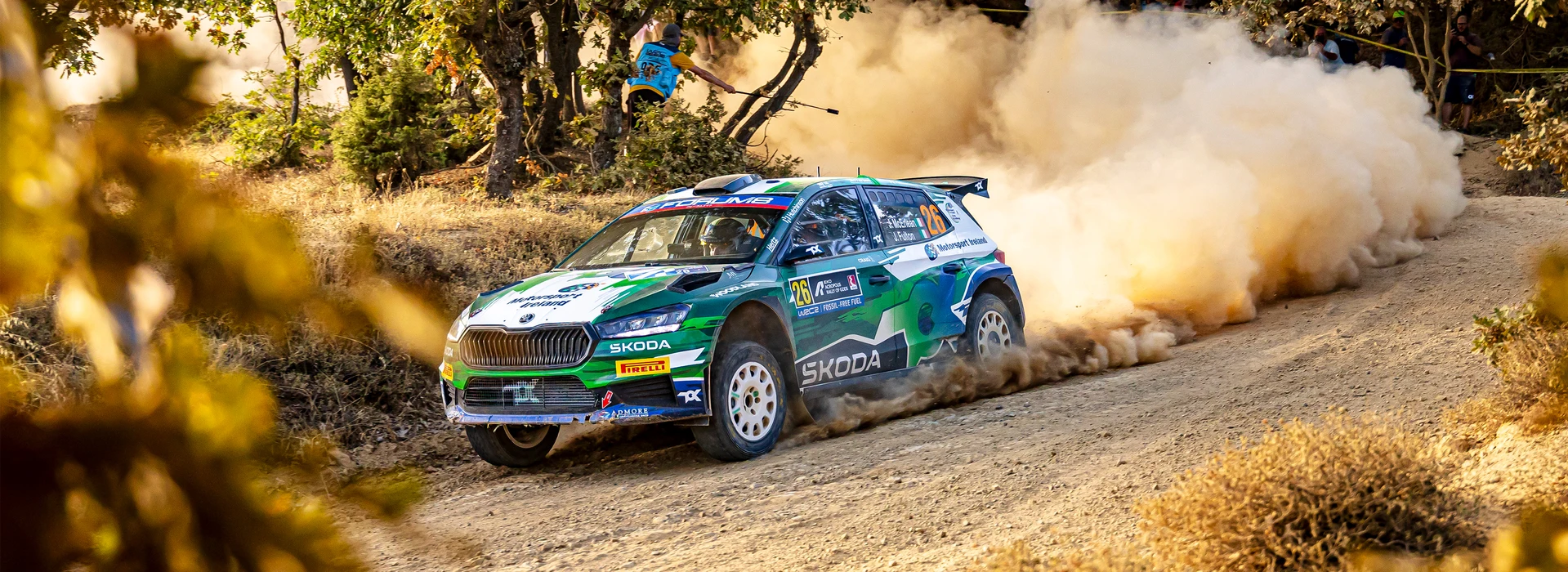 Une voiture de rallye Skoda Fabia verte et blanche avec le numéro 26 dérape sur une piste de terre, soulevant un grand nuage de poussière, avec des spectateurs et des arbres en arrière-plan.