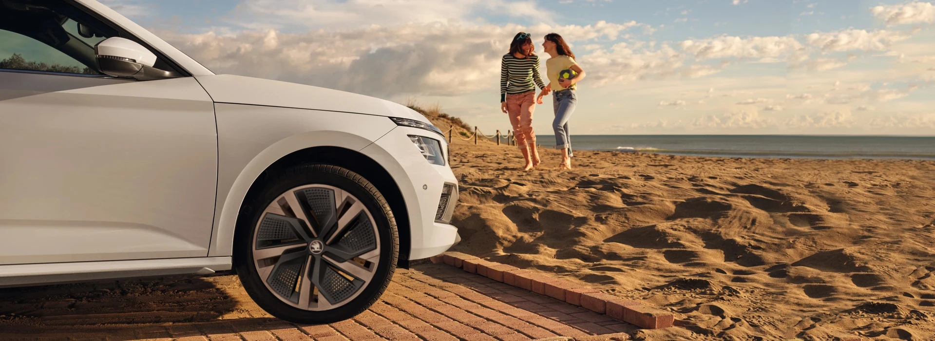Vista di un veicolo Škoda parcheggiato in spiaggia e due ragazze che si tengono per mano.