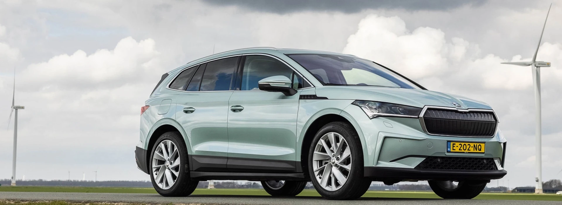 Een Skoda Enyaq rijdt op een weg door landelijk gebied, op de achtergrond een windmolen