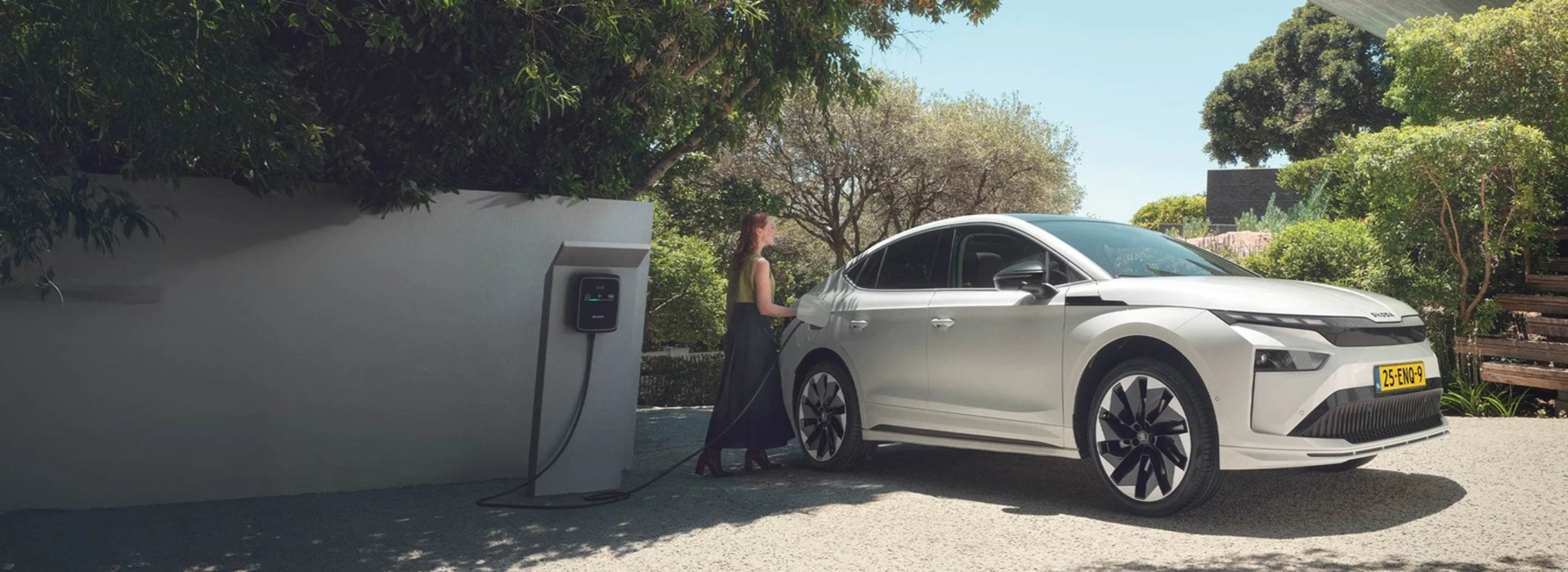 Een vrouw steekt een oplaadstekker in een witte Skoda die geparkeerd staat op een oprit in natuurrijk gebied