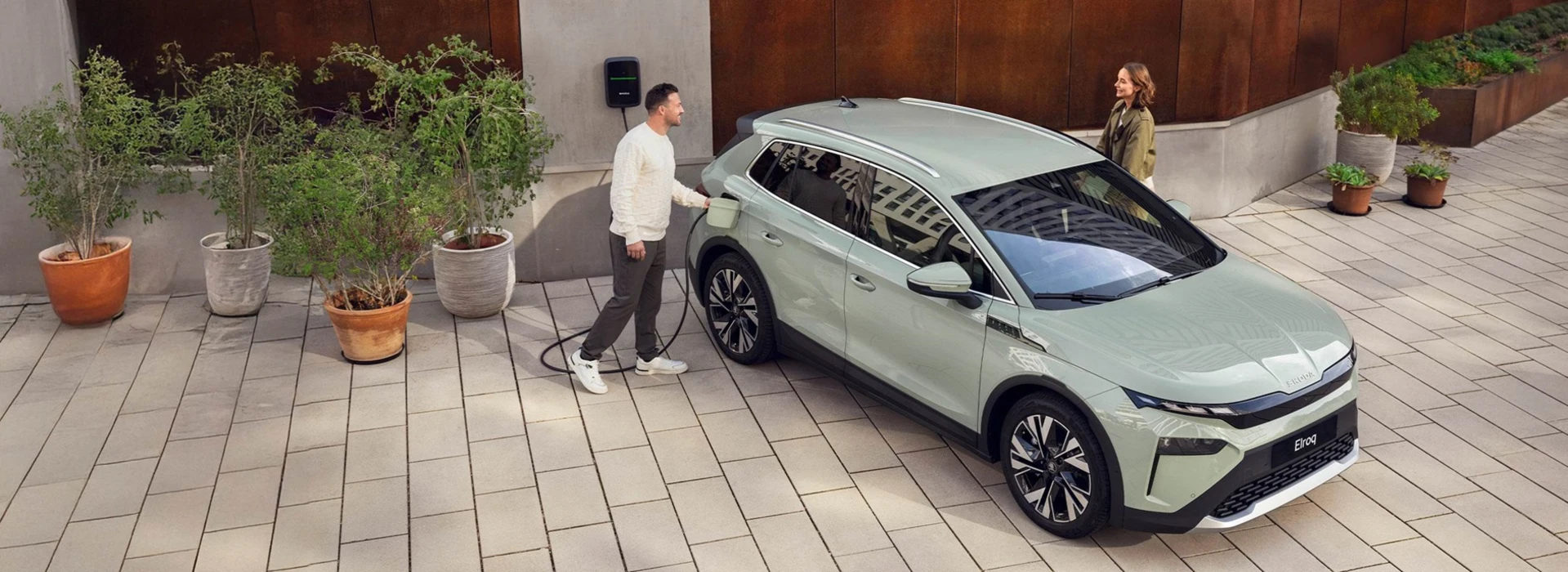 Een man haalt een stekker uit een Skoda elroq die geparkeerd staat op een oprit, een vrouw wil aan de ander kant van de auto instappen