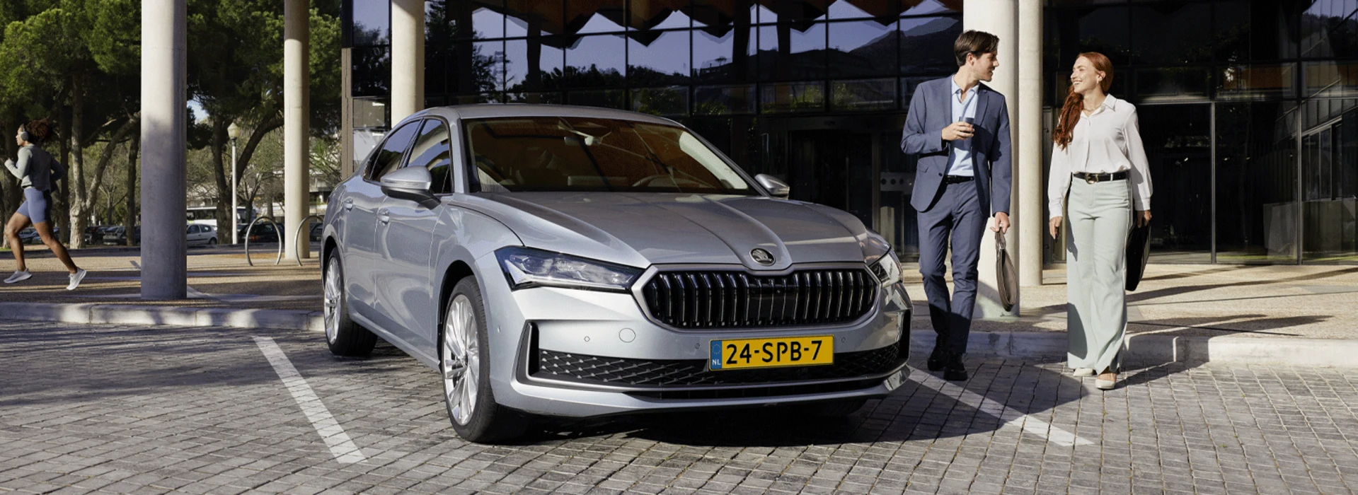 Een stel of een paar collega's lopen naast een grijze Skoda Superb die geparkeerd staat op een bedrijventerrein