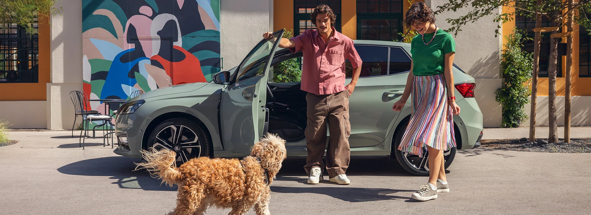 Ett ungt resesällskap, kvinna, man och hund, framför den rymliga småstadsbilen Fabia i snygga Timiano Green.